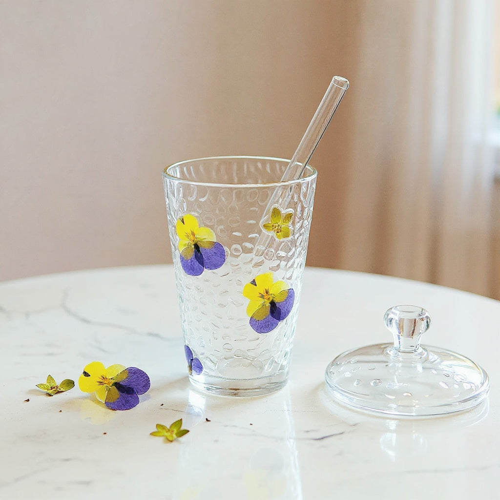 Transparent glass cup with real dried flowers embedded on surface, glass lid and straw, surrounded by scattered petals on a marble table.