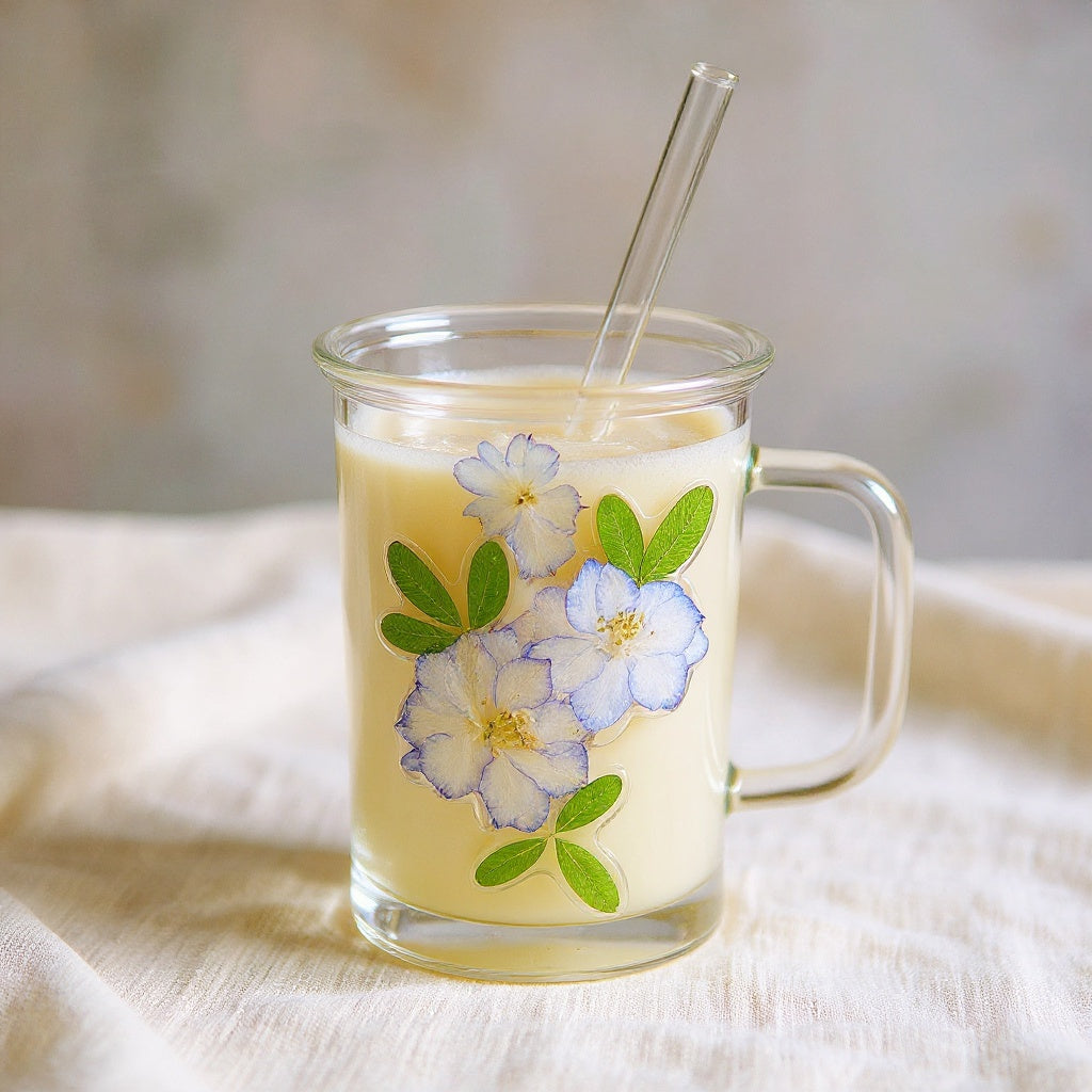 Square-profile botanical glass drinkware with preserved flowers, glass lid, and straw on a marble surface in soft natural light.