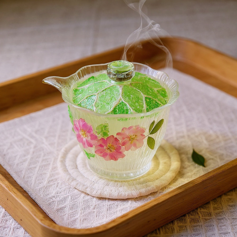 A delicate green-tinted glass tea cup with raised lotus petals and embedded dried flowers on a linen cloth, steam rising gently in warm natural light.