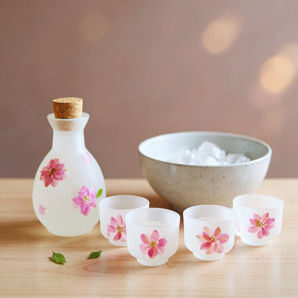 Soft matte frosted glass sake set with embedded cherry blossom petals on a pale wood surface, soft bokeh background.