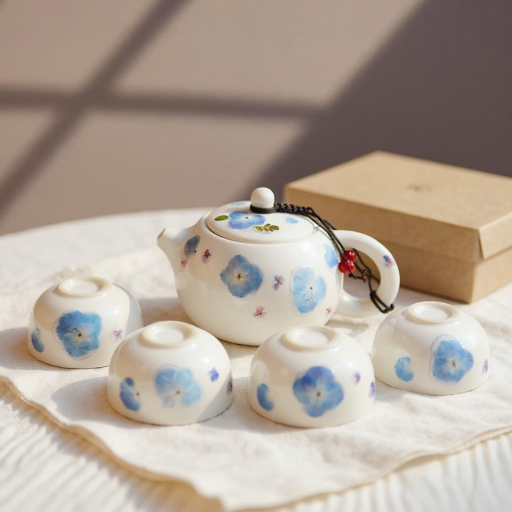 Luminous white porcelain tea set with real pressed blue hydrangea relief on a linen surface, gift box in soft natural light.