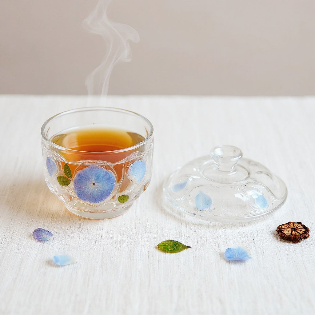 A delicate transparent glass teacup with saucer and lid, embedded with blue-purple hydrangea petals, surrounded by scattered dried petals on a neutral background, with steam rising softly.