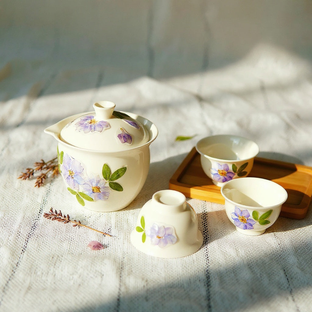 A semi-translucent warm white porcelain gaiwan and two teacups with embedded dried flower petals in soft pinks and lavenders, set on a linen surface with scattered botanical props, wide composition with negative space.