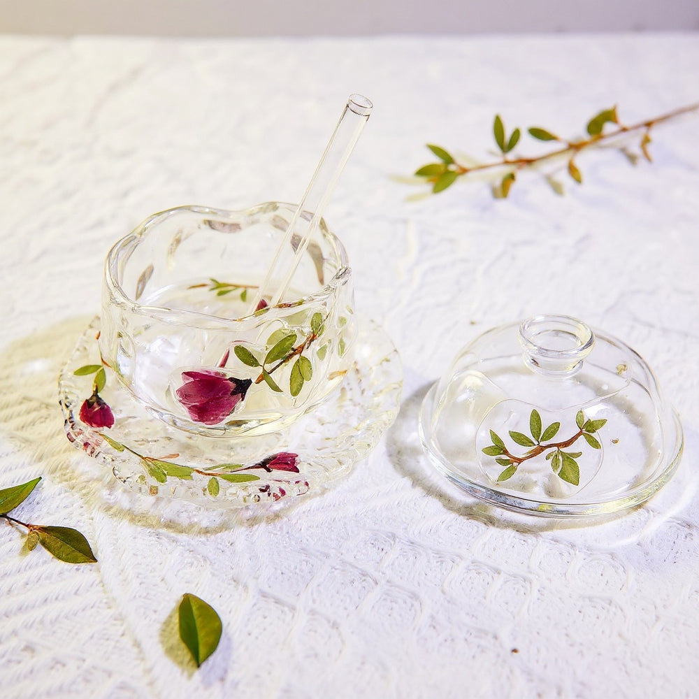 Transparent borosilicate glass teacup with embedded roses, saucer, glass straw, and lid on marble with dried botanicals, wide editorial shot.
