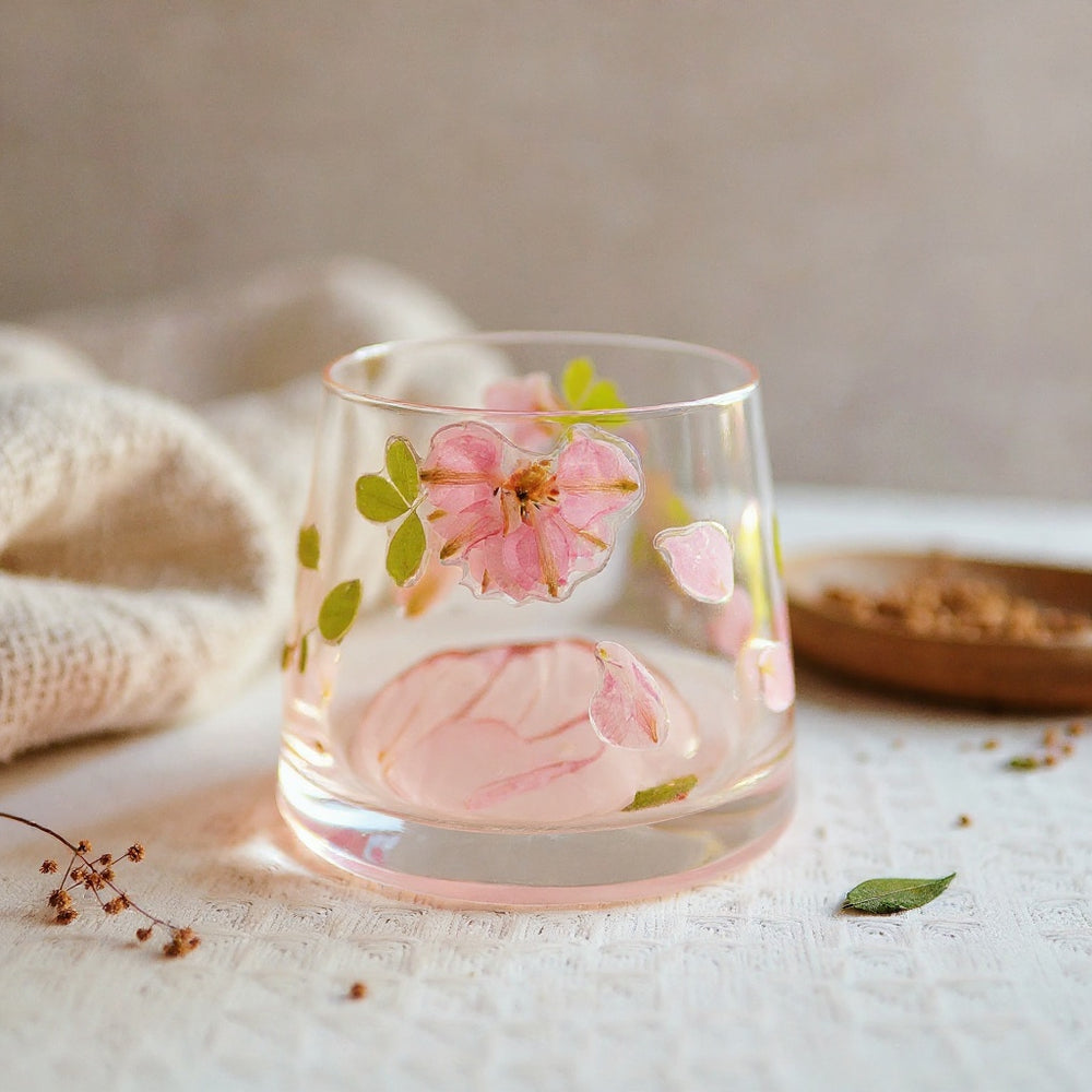 Cylindrical glass tumbler with real preserved flowers embedded on the surface, styled with natural materials on a neutral background, wide composition with soft lighting.