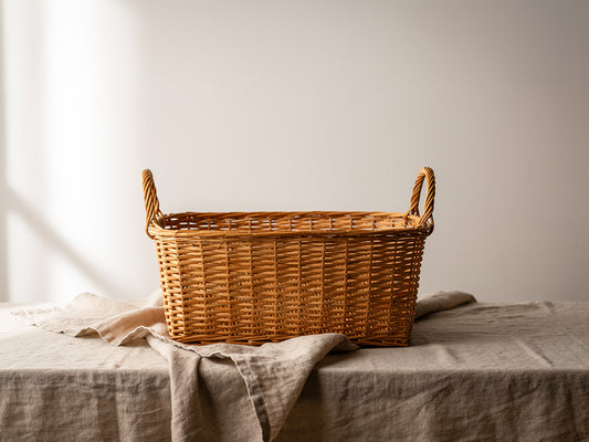 Rectangular handwoven rattan basket in honey-tone with dual arched handles, styled on a neutral background with natural materials and soft lighting in a wide editorial composition.