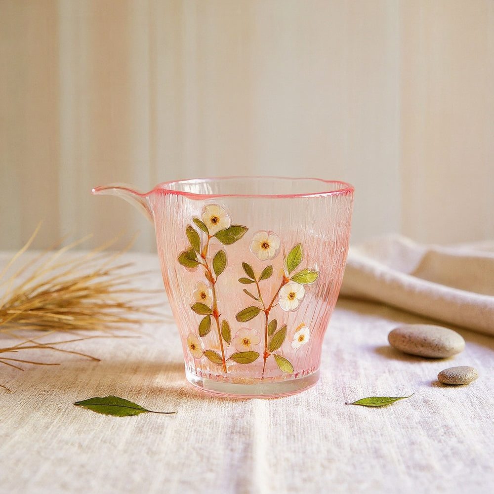A clear glass tumbler with embedded real blush, cream, and green flower petals, set on natural linen with soft lighting.