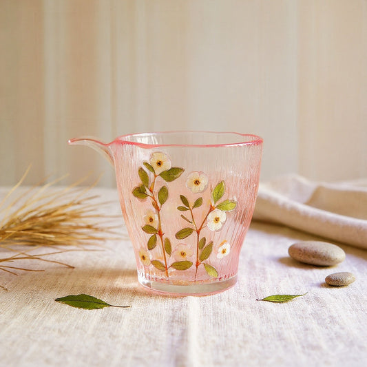 A clear glass tumbler with embedded real blush, cream, and green flower petals, set on natural linen with soft lighting.