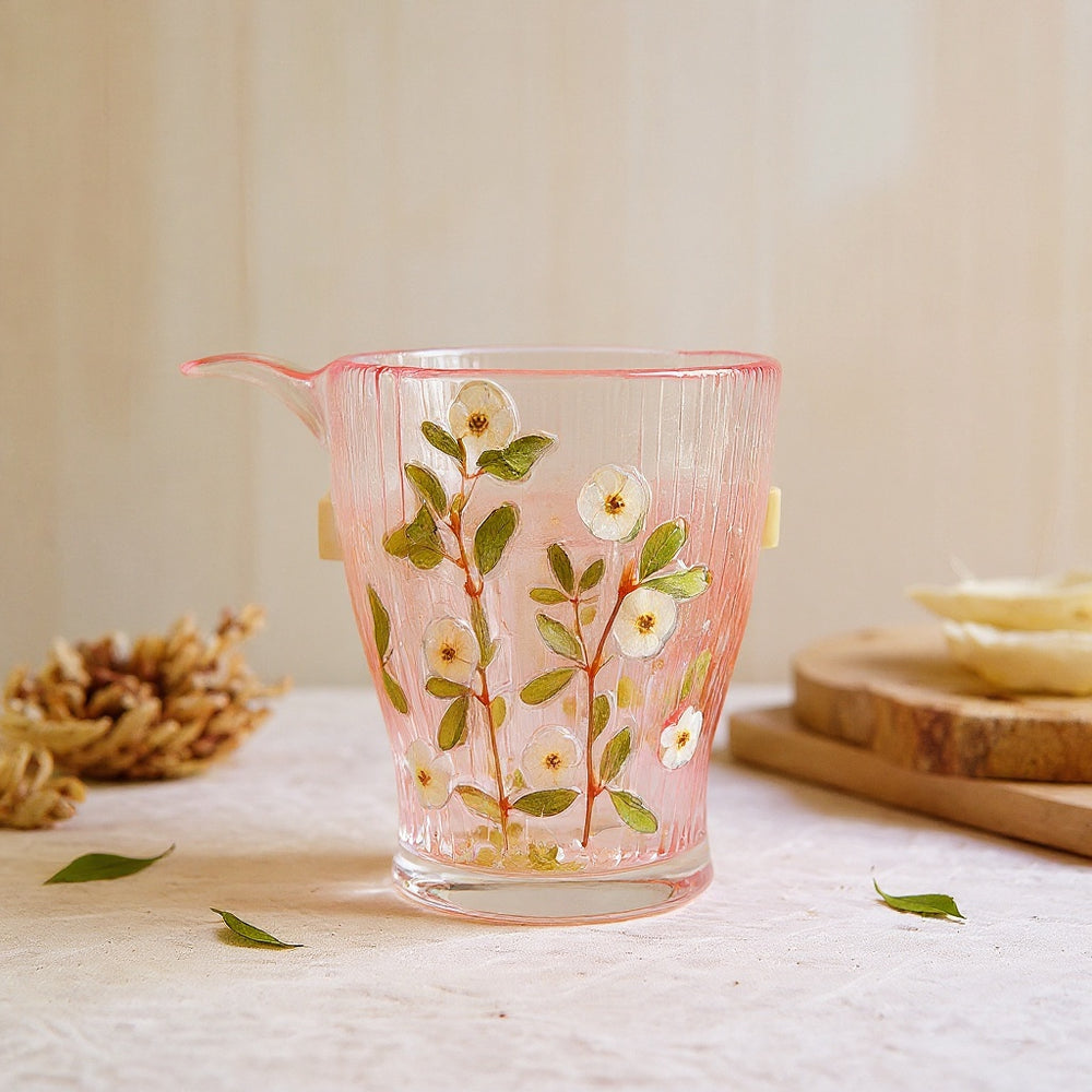 Cylindrical glass tumbler with embedded real preserved flowers and leaves, set against a neutral background with natural props, soft editorial lighting.