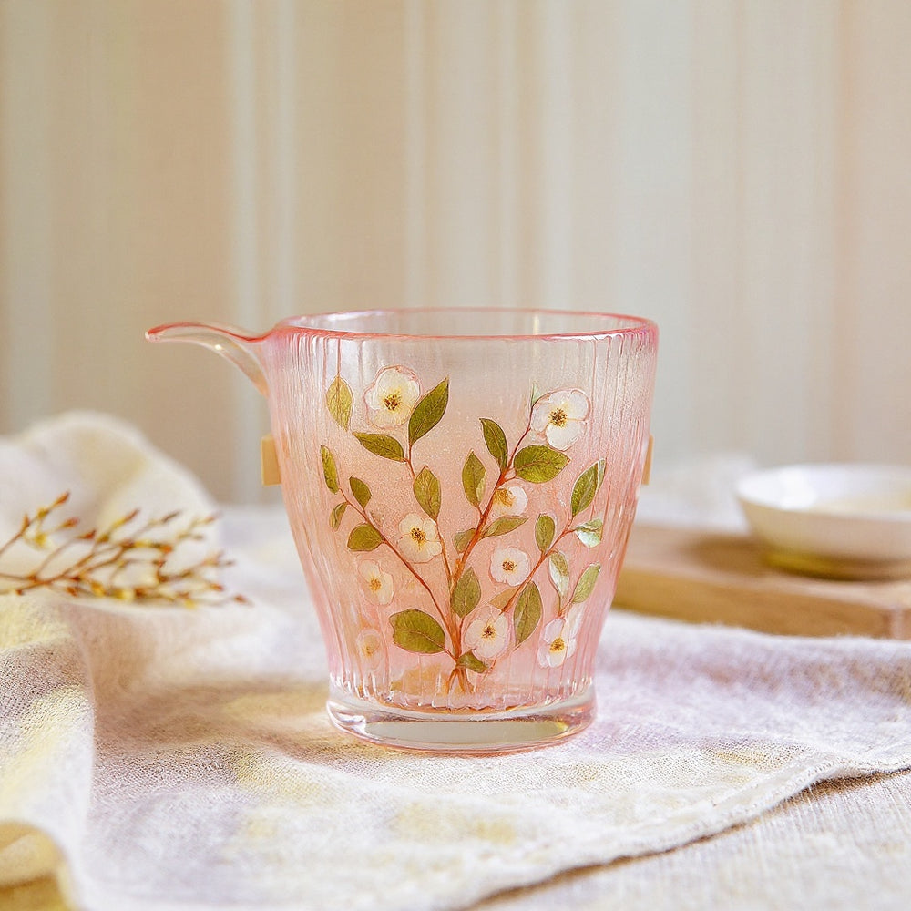 Cylindrical glass tumbler with real preserved flowers on a neutral textured surface with botanical props, wide editorial composition.