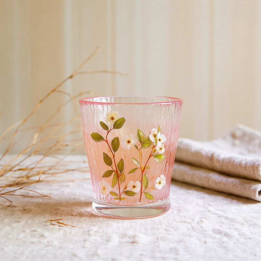 A cylindrical glass tumbler with embedded preserved flowers sits on a linen cloth, surrounded by soft natural elements.