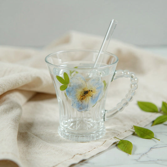 Clear glass cup with dried flowers embedded on the surface, beaded handle, frosted lid, and glass straw, on a linen cloth with loose dried flowers, wide composition.