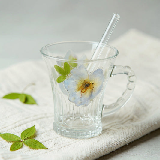 Clear glass tea cup with embedded pink dried flowers, beaded handle, and straw, on linen with scattered dried botanicals in soft natural light.