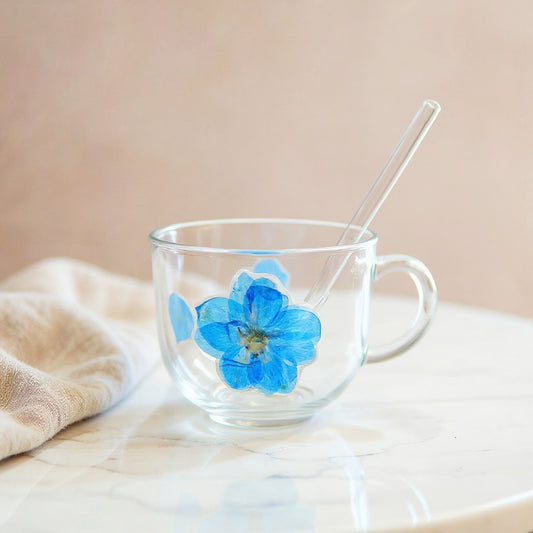Hero image of a floral glass cup with lid and straw on a white marble surface, soft natural lighting, minimalist setting.