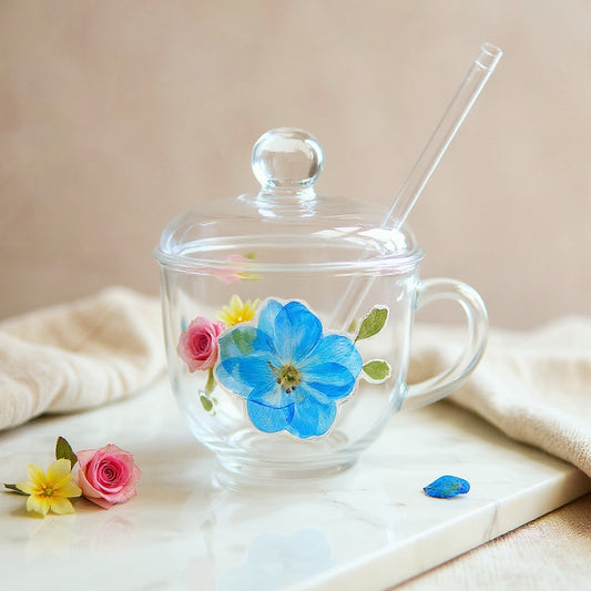 Clear glass cup with 3D floral relief of pink roses, yellow and blue blooms, with glass lid and straw, on neutral linen surface.
