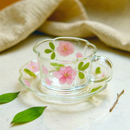 Handcrafted glass cup and saucer with embedded pink flowers on a cream marble surface with linen cloth and twig.