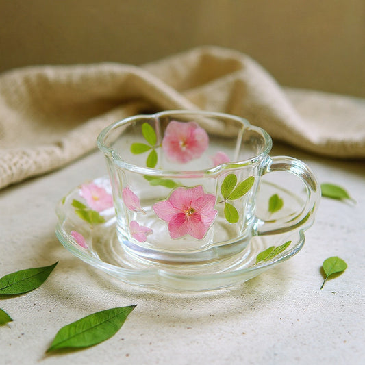 Clear high-borosilicate glass cup and saucer with handcrafted botanical relief, real pink petals, on neutral backdrop with soft shadows.