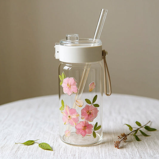 Elegant artisan flower glass tumbler with dried pink petals, dust-proof lid and straw, on a linen surface with loose botanicals in soft natural light.