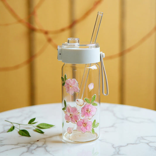 A clear glass tumbler with real dried flowers on its textured exterior, with a frosted lid and a glass straw, on a marble surface surrounded by loose dried botanicals, in soft natural light.