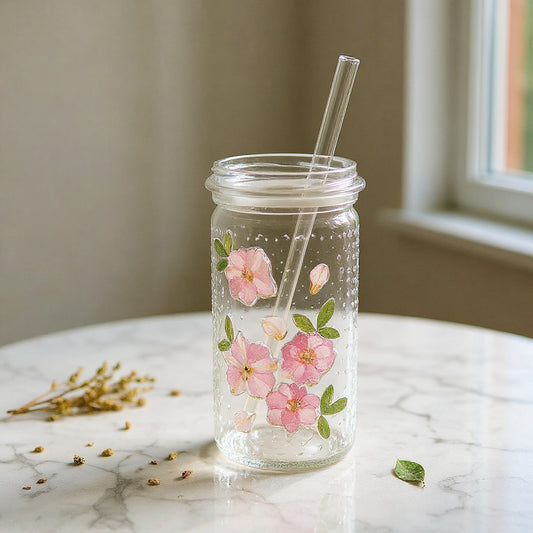 Clear glass tumbler with dried flowers, lid, and straw on marble with loose botanicals.