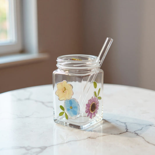 Short square glass with embedded pastel flowers, lid, and straw on marble, soft lighting.