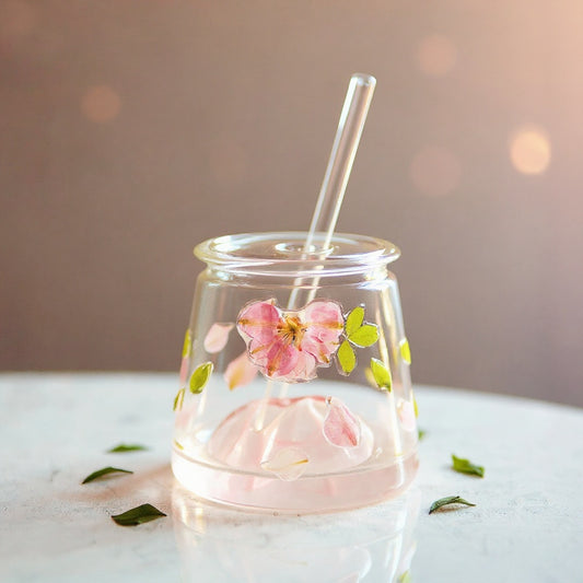 A clear glass tumbler with preserved flowers and leaves on the exterior, lid and straw, on marble with pink bokeh background