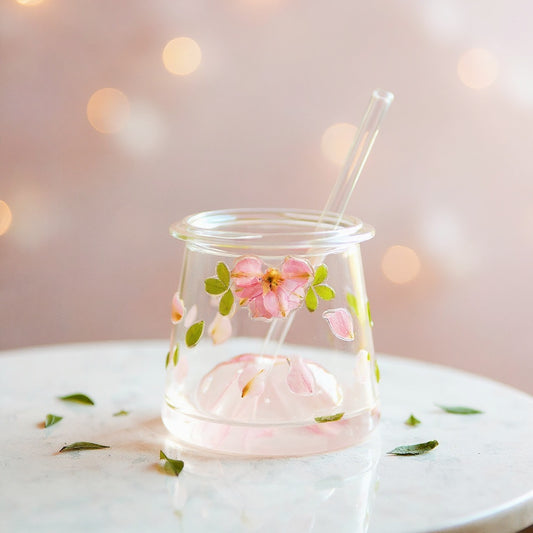 A clear glass tumbler with three-dimensional preserved flowers and a glass straw, set on a white marble surface against a softly blurred pastel background.
