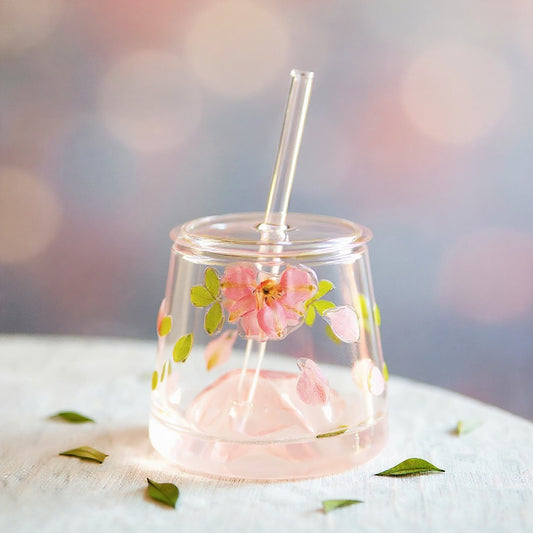 A clear glass tumbler with preserved flower petals and leaves in 3D relief, glass lid and straw, on a light surface with soft blurred background.