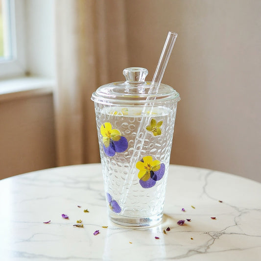 Clear glass cup with embedded dried flowers and straw on a marble surface with scattered petals