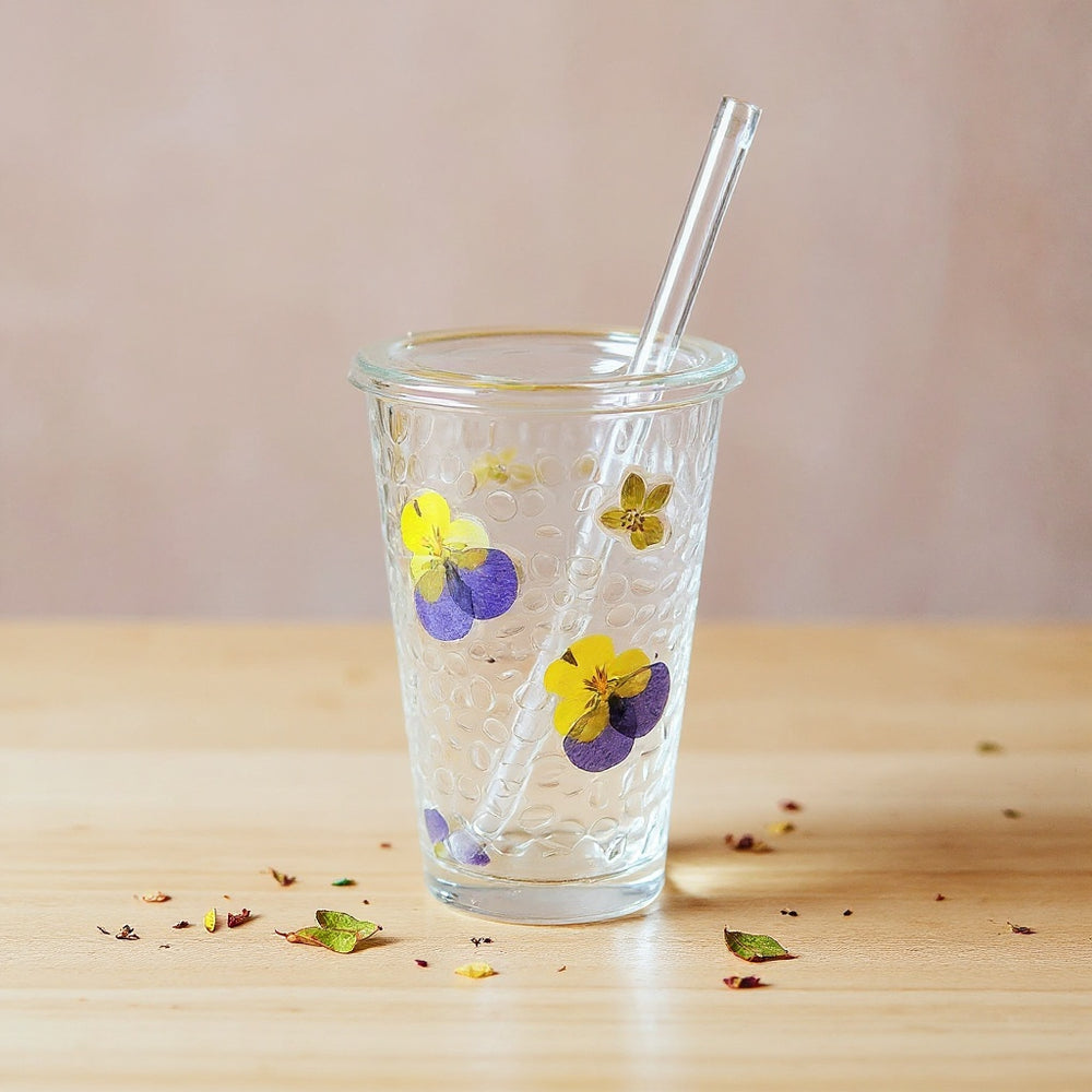 Artisan glass cup with embedded dried flowers on a wooden surface, soft lighting, scattered petals.