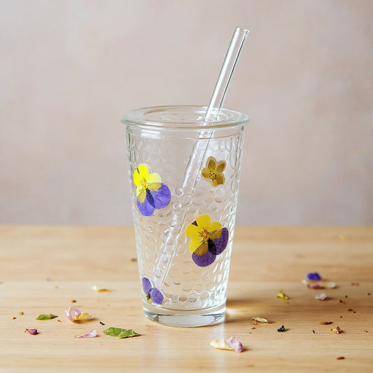 Handmade preserved flower drinking glass on wooden surface with scattered dried petals, soft natural lighting.