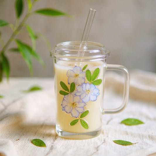 Square heat-resistant glass cup with preserved flowers in raised relief, pastel drink, and matching lid, on a soft-lit marble surface.