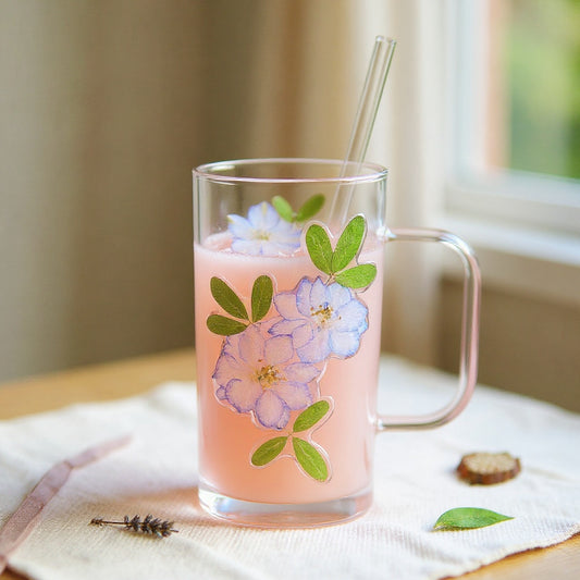 Square-profile glass cup with embedded dried flowers, pastel drink, glass lid, and straw, on a linen surface with soft natural lighting and botanical accents.