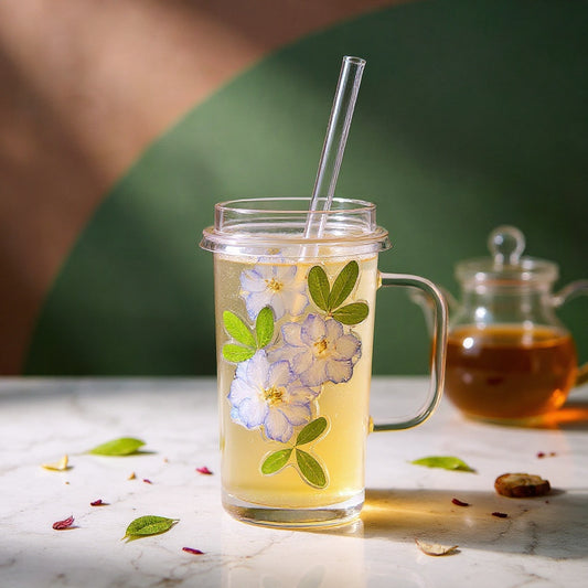 Square glass cup with lid and straw, embedded with dried flowers, on marble with a blush and sage gradient background, scattered rose petals and glass teapot.