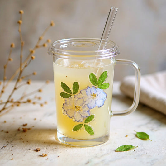 Square glass cup with embedded botanicals and glass straw on a natural surface with soft lighting.