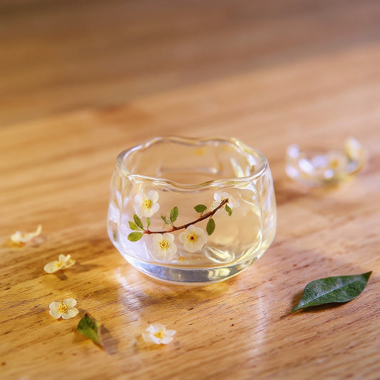 A handcrafted clear glass tea cup with preserved white plum blossoms and green leaves, on a wooden surface with scattered petals, soft natural light.