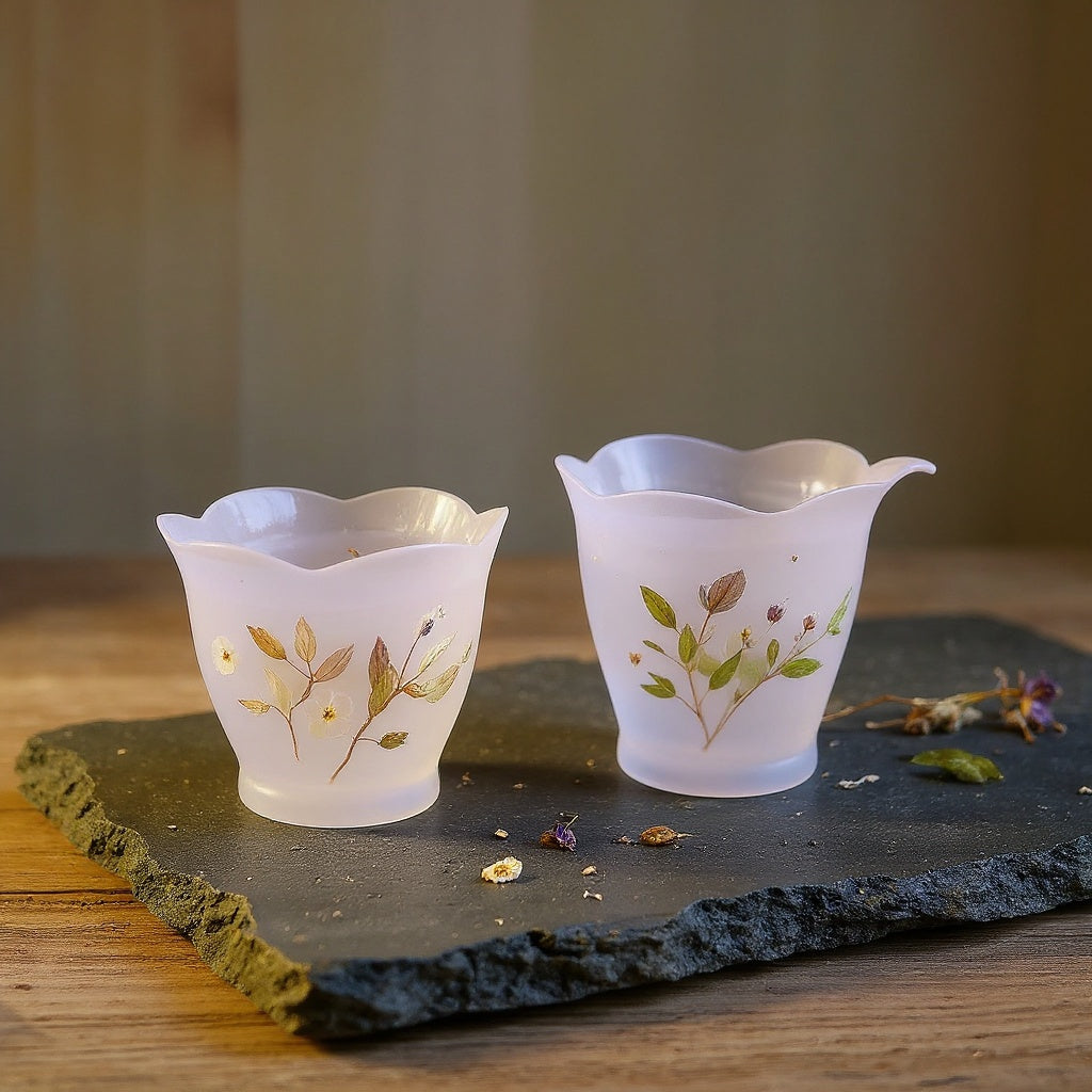 Frosted translucent purple lotus-petal glass cup with embedded dried flowers and foliage on raw wood with loose dried flowers, soft natural lighting.