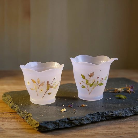 Frosted translucent purple lotus-petal glass cup with embedded dried flowers and foliage on raw wood with loose dried flowers, soft natural lighting.