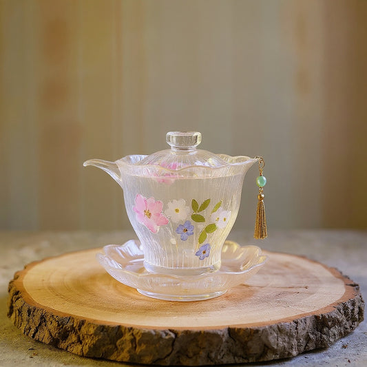 Luxury translucent lotus-shaped glass teacup with embedded real pressed flowers and jeweled tassel on a raw wood surface, soft bokeh background, editorial botanical aesthetic