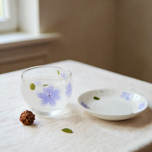 Frosted glass teacup with embedded hydrangea petals on a linen surface with saucer and dried botanicals, soft natural lighting.