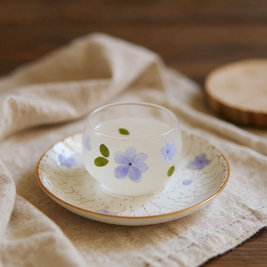 Small frosted glass teacup with blue and purple hydrangea petals embedded in resin, on a textured floral saucer, with soft natural light on a neutral background, wide composition.