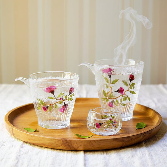 A transparent hammered glass gaiwan and cups with real pink rose petals and green leaves sealed on the surface, arranged on a wooden tray with steam rising, against a soft linen background.