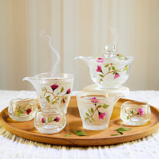 A lotus shaped gaiwan set made of transparent ice-ripple hammered glass with pink rose petals and green leaves in 3D relief, arranged on a wooden tea tray with steam, against a white linen background.