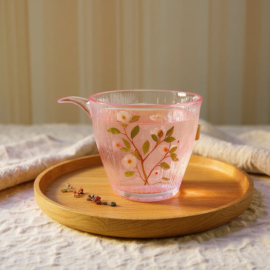 Luxury glass tea cup with embedded dried plum blossom petals and leaves on a wooden tray with linen, soft natural lighting.
