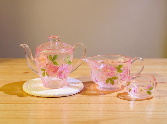 Handmade glass teapot and teacup set with real cherry blossom petals embedded, soft pink tones, on a wooden surface in warm natural light.