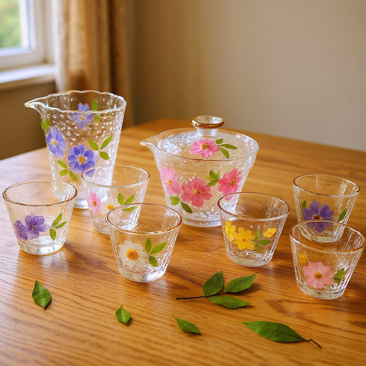 Complete 8-piece modern gongfu tea set in transparent hammered glass with embedded dried flowers, arranged on a wood surface in soft natural light.