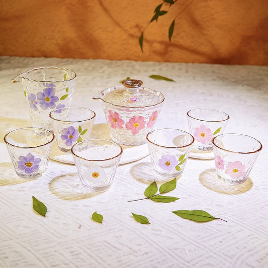 A set of 8 transparent hammered glass tea cups with embedded real pressed flowers and gold rims, elegantly arranged on a marble surface with soft natural light, wide editorial composition.