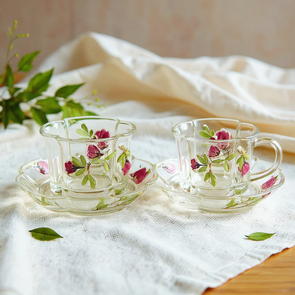 A delicate clear glass teacup and saucer with embedded dried flower petals and leaves, styled on a linen tablecloth with soft natural lighting.