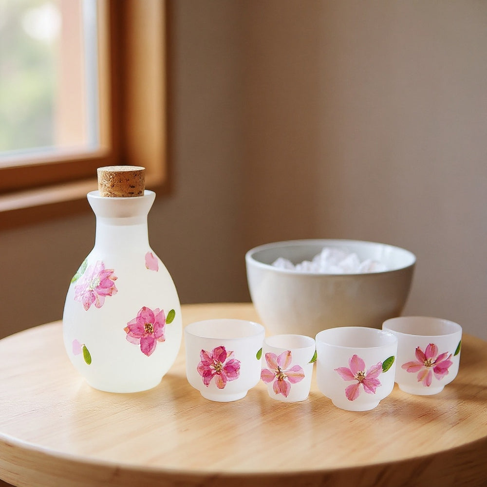 Soft matte frosted glass sake carafe set with pressed cherry blossoms and ice-filled chiller bowl on pale wood, warm natural light, bokeh background.