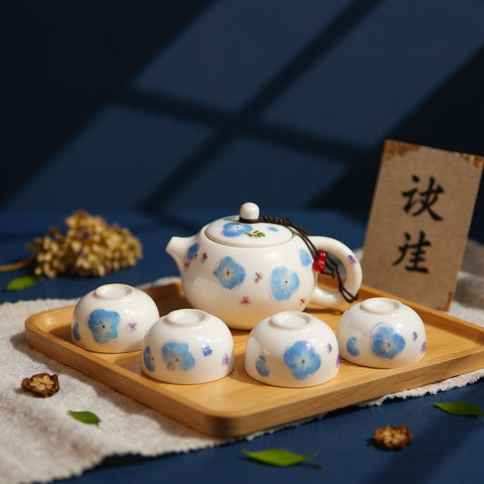 White porcelain teapot and teacups with real pressed blue hydrangea petals on a linen runner with dried blooms against a blue gradient background.